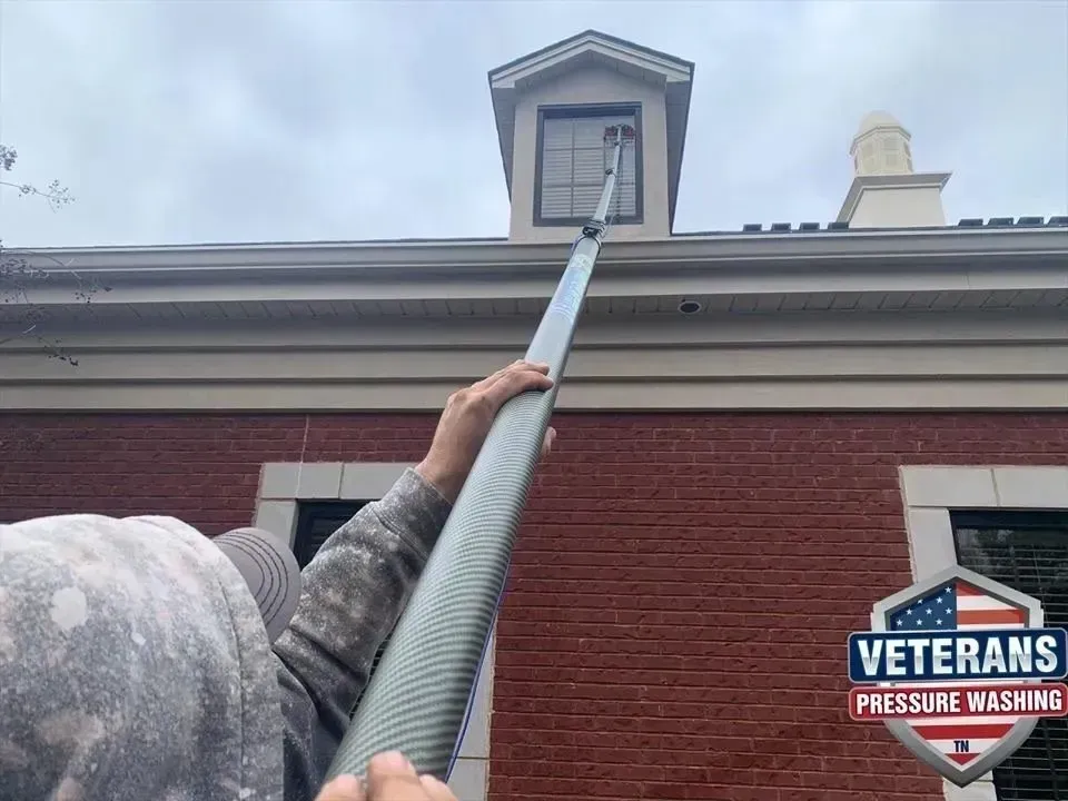 Person using an extendable pole to clean a window on a brick house with cloudy sky.