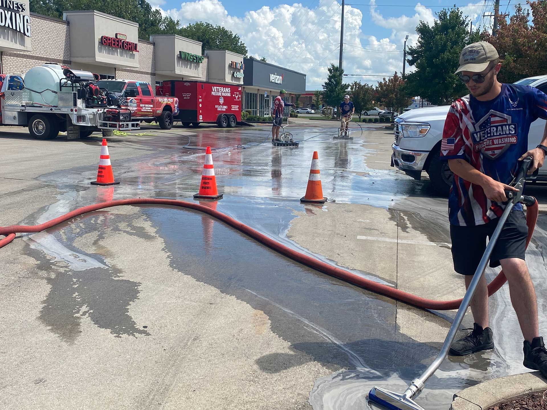 Man washing a wet parking lot with a long brush and hose, orange traffic cones, fire trucks in the background.