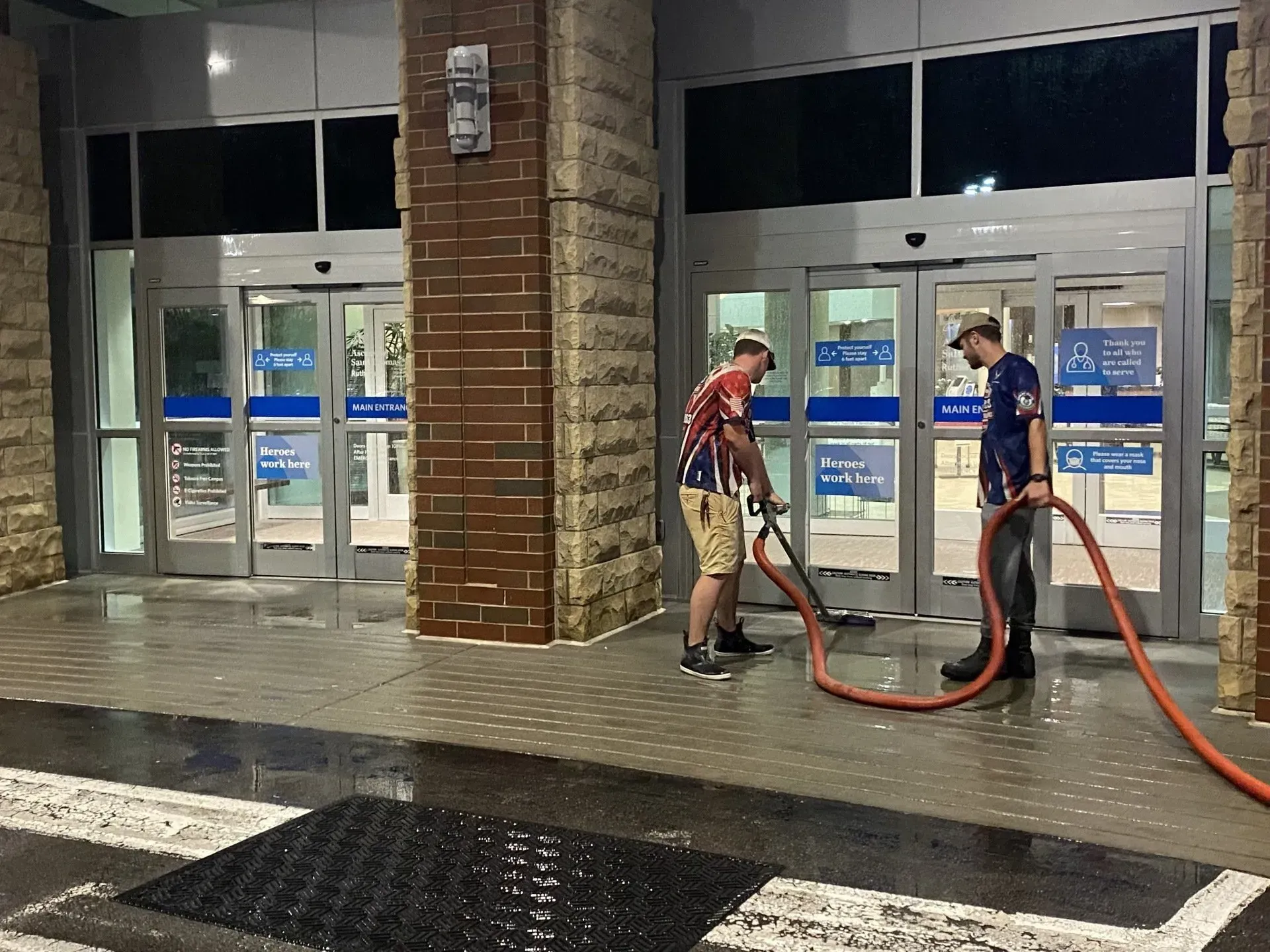 Two people washing the entrance of a building with a hose. Brick building with automatic doors.