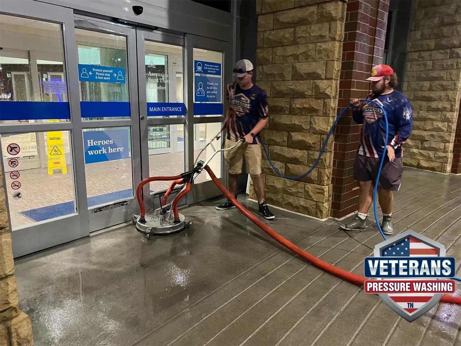 Two veterans power washing a store entrance. One holds the hose, the other guides the washer.