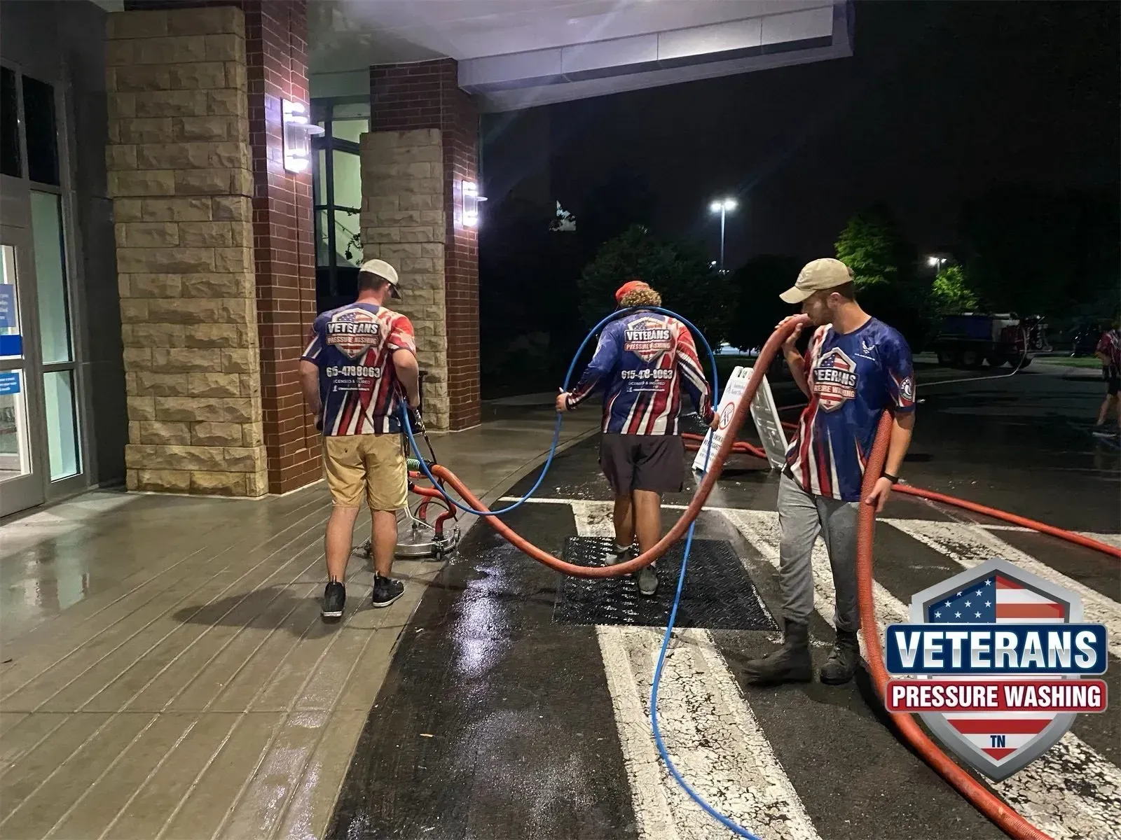 Three people pressure washing a commercial building exterior at night.