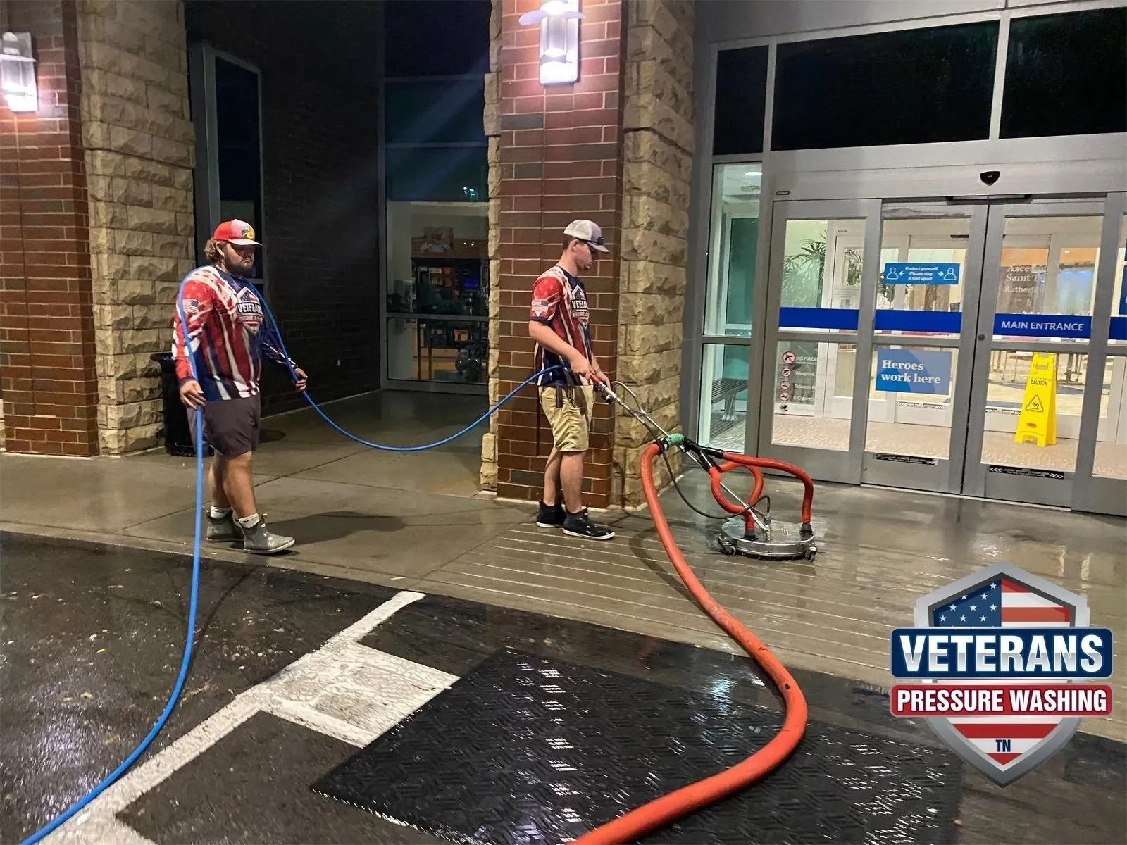 Two men pressure washing a store entrance at night. One operates the cleaner, the other manages the hose.