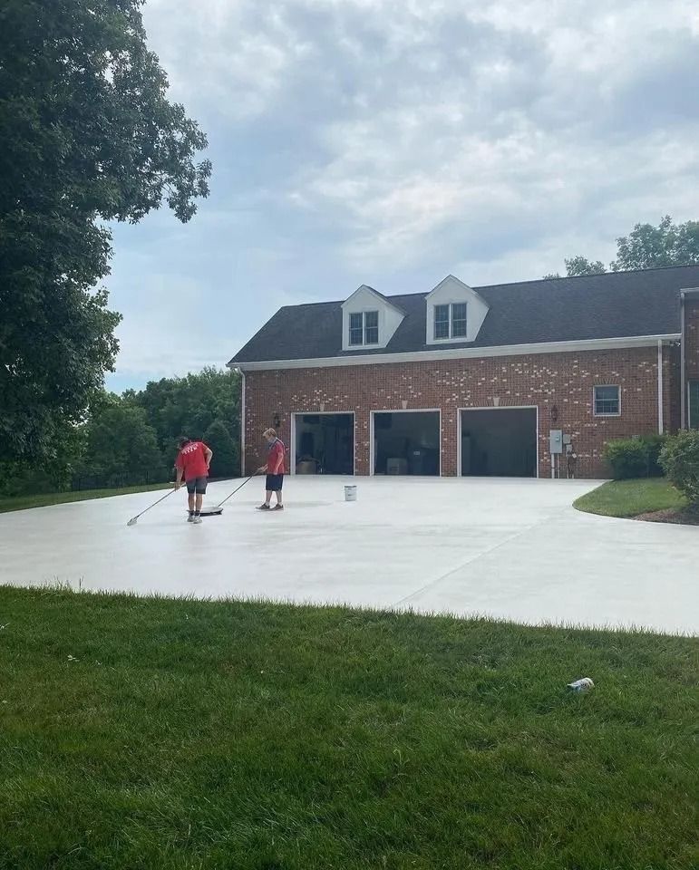 Two people painting a light grey driveway in front of a brick house with a three-car garage.