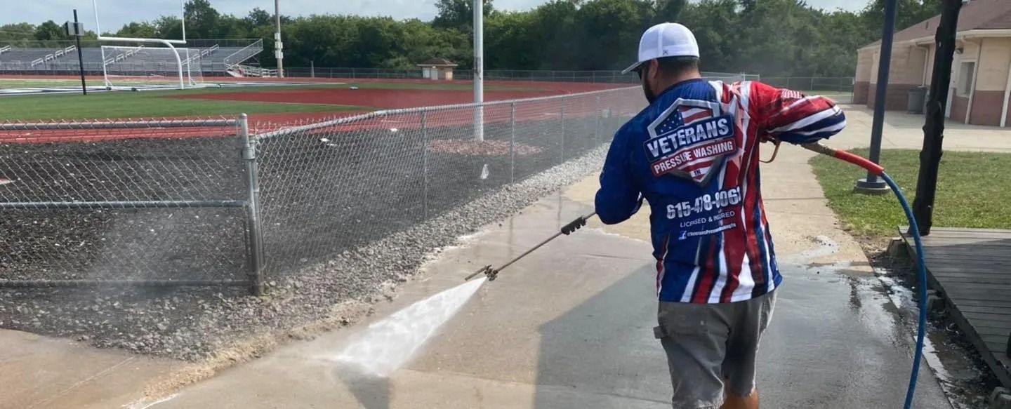 A man power washing a concrete walkway next to a chain-link fence and a sports field.