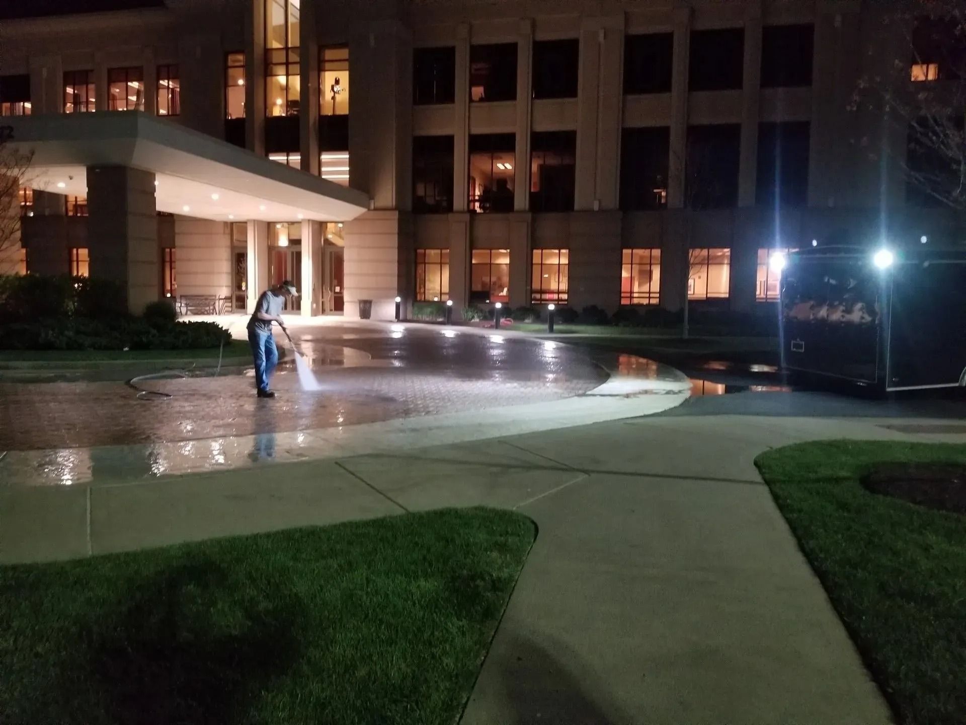Person pressure washing a paved entrance to a large building at night, a black van parked nearby.