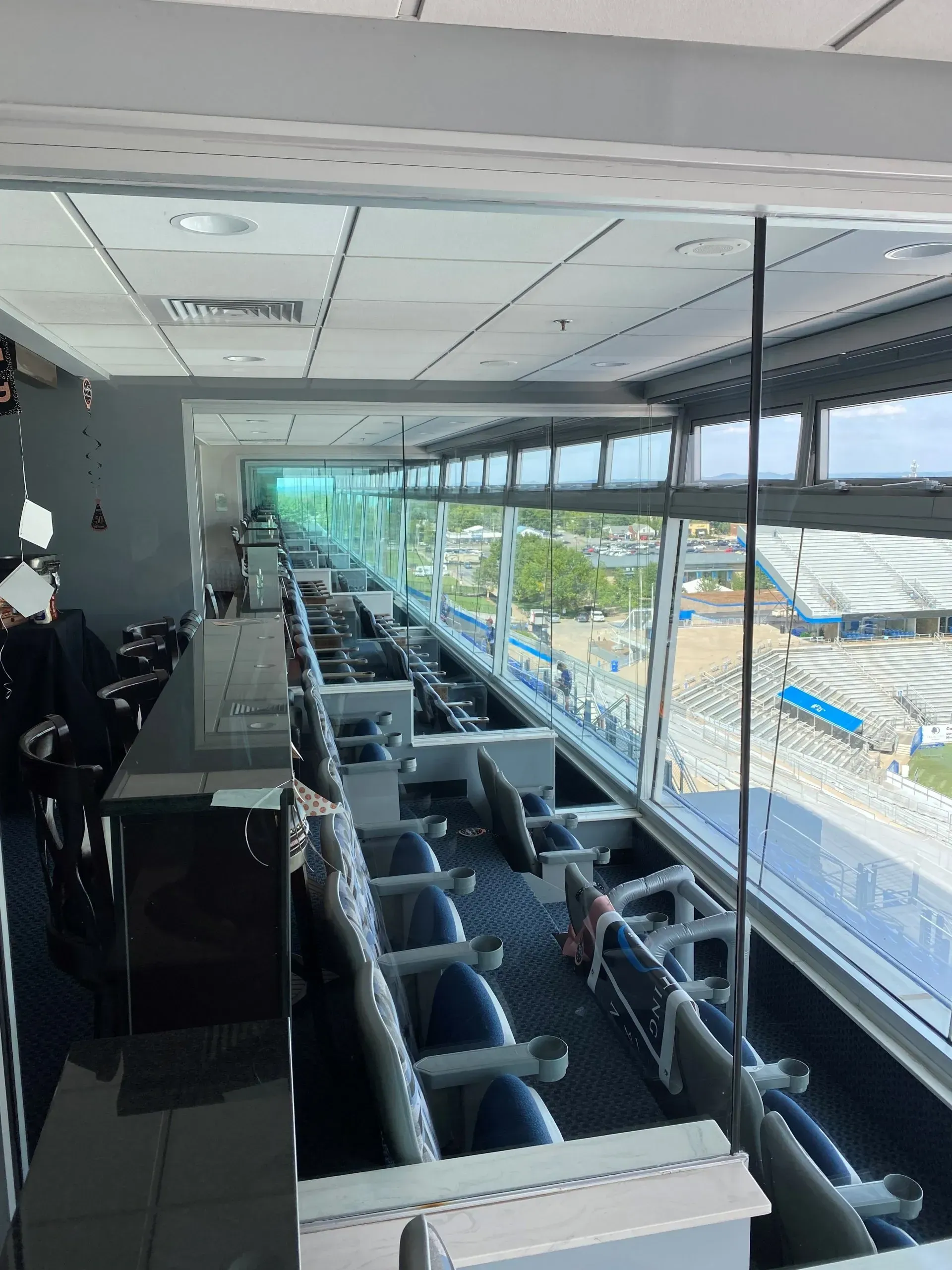 Long, empty stadium suite with rows of seats, overlooking a sunny stadium with blue and white seating.