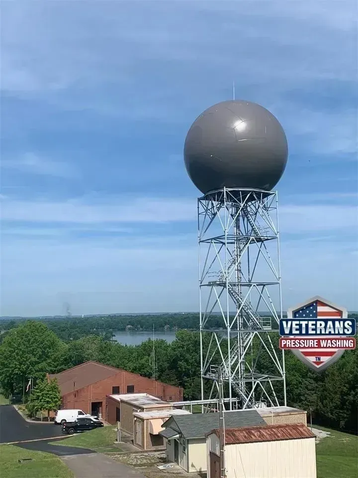 A large gray weather radar tower with a round dome, green trees, and a blue sky.