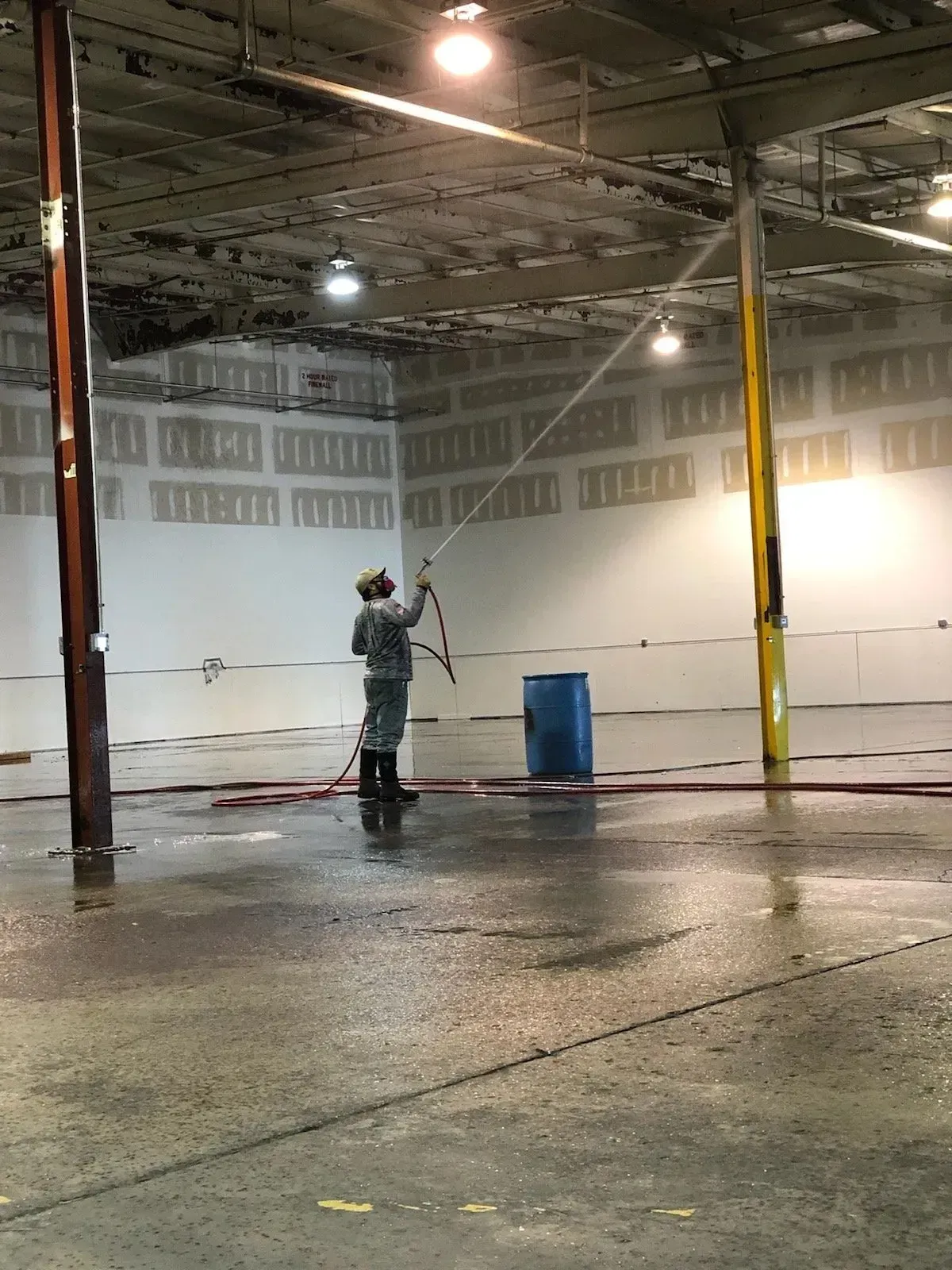 Person spraying a large industrial space. Gray suit, protective gear. Water on floor. Blue barrel, metal poles.
