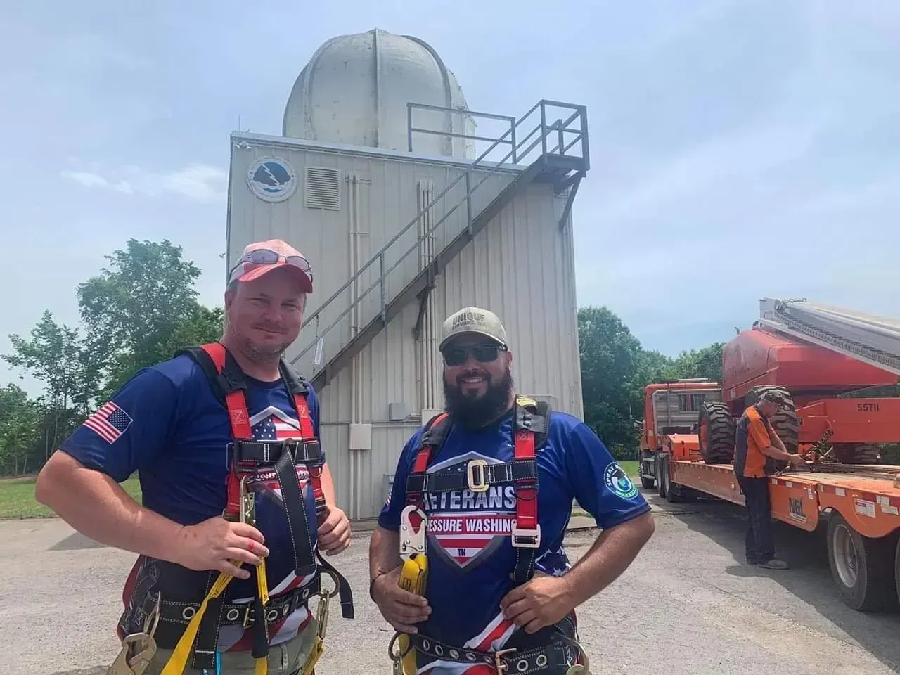 Two men in safety harnesses stand in front of an observatory with a truck and trailer in the background.