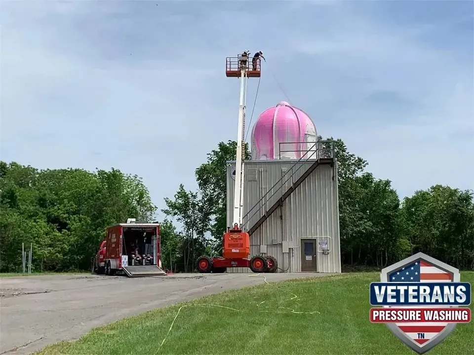 Two people on a lift cleaning a pink dome, next to a utility building. Veterans pressure washing van nearby.