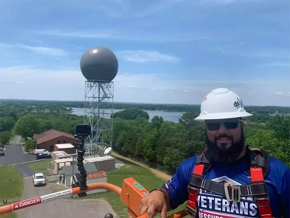 Man in hard hat and harness at radar tower with lake backdrop.