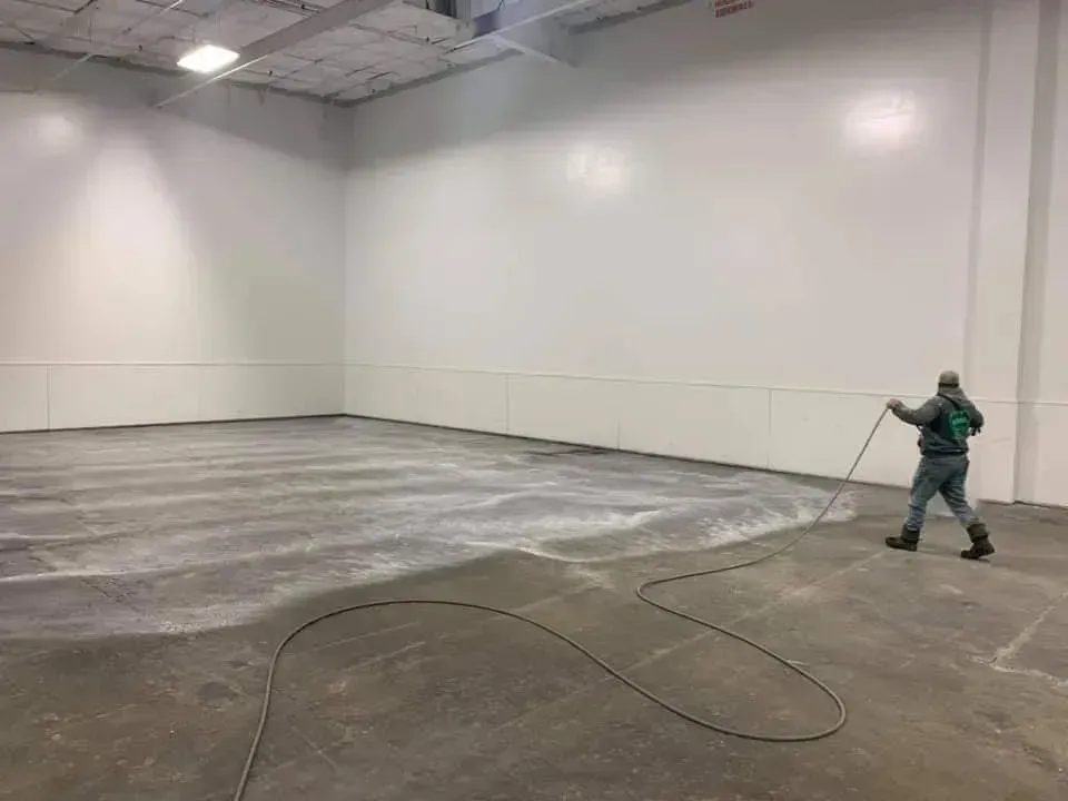 Person spraying cleaner on a large concrete floor in a white-walled warehouse.