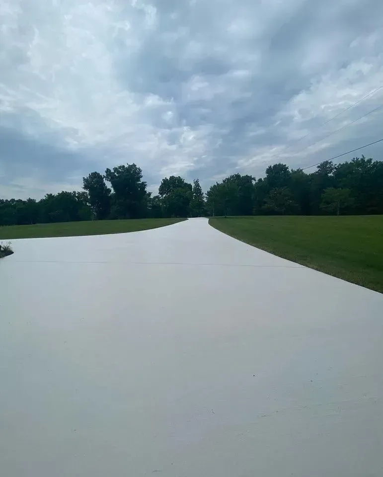 A wide, light-colored concrete driveway leads into the distance under a cloudy sky. Green grass borders it.