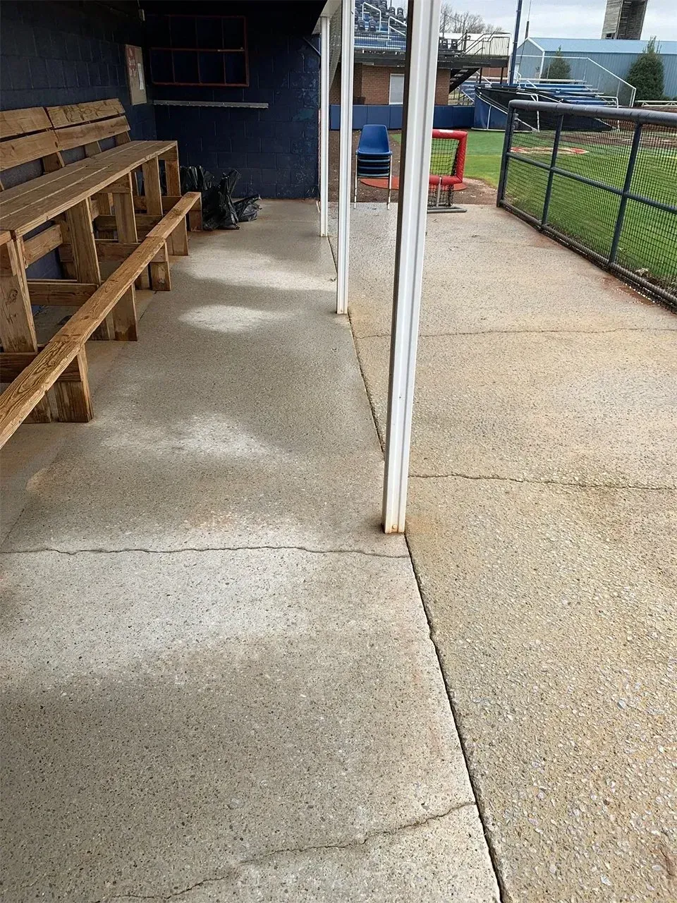 A concrete dugout with wooden benches, white support poles, and a view of a baseball field.