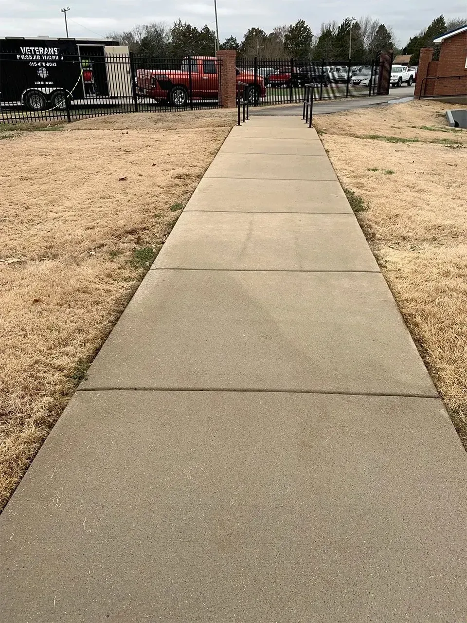 Concrete sidewalk leading toward a gated area with red truck and people; brown grass on either side.