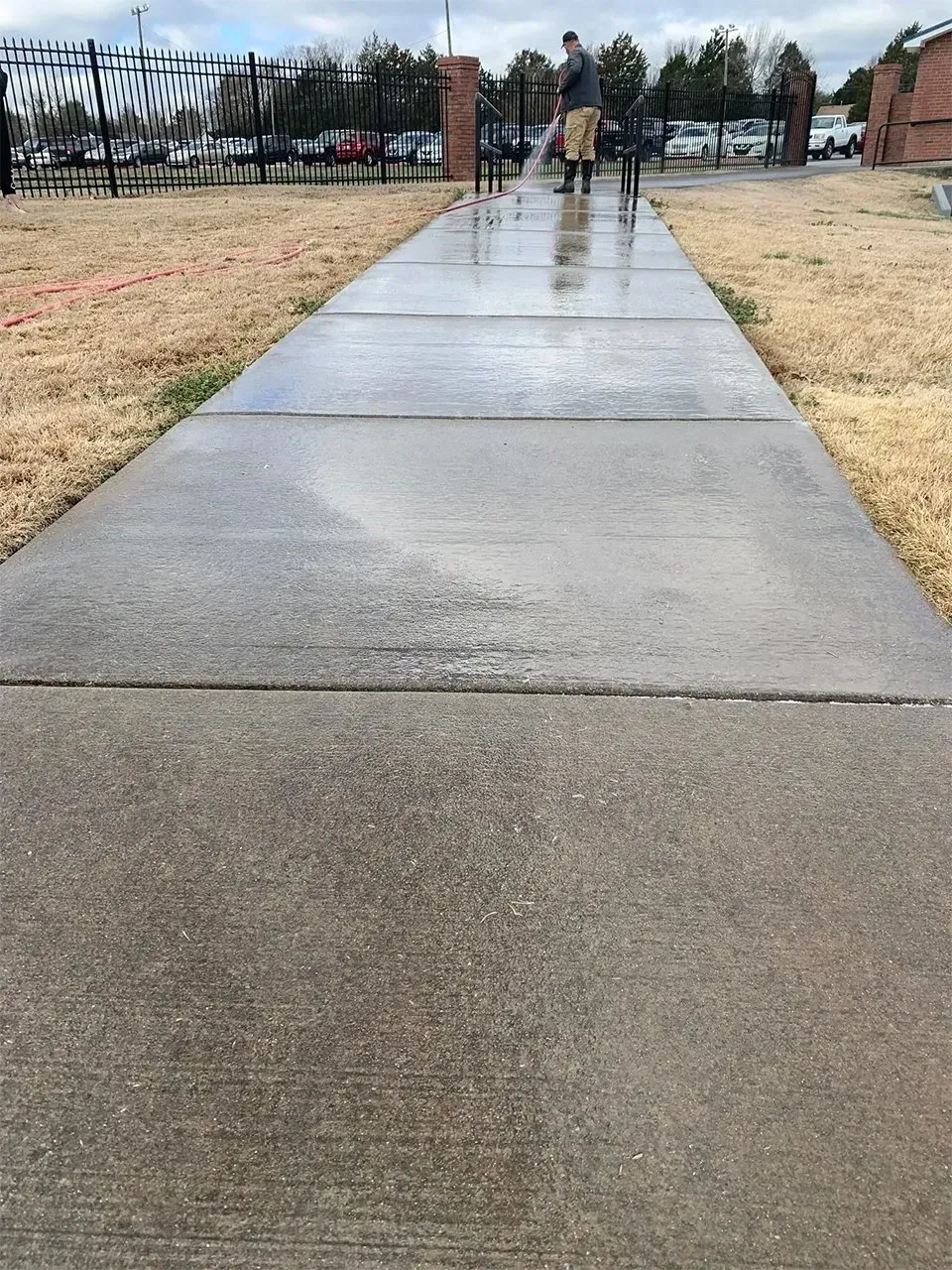 Man power washes a concrete sidewalk, damp and reflective, in front of a building with a fence.