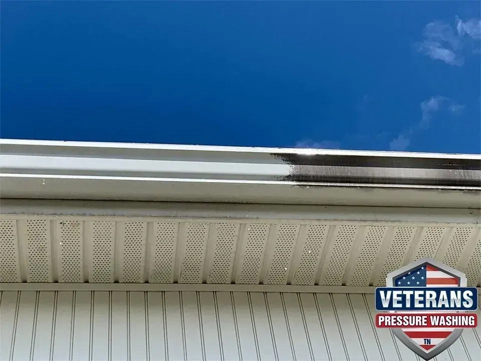 White gutter being pressure washed, removing black streaks against a blue sky.