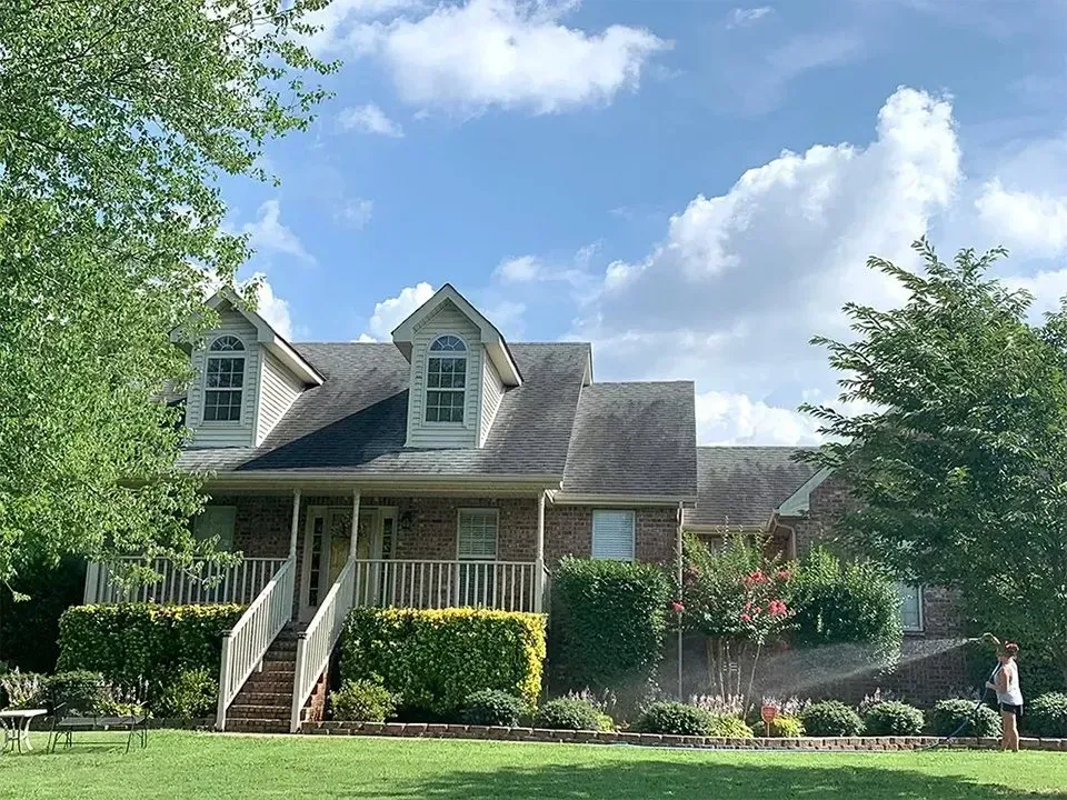 Two-story brick house with dormers, porch, and lush landscaping under a partly cloudy sky.