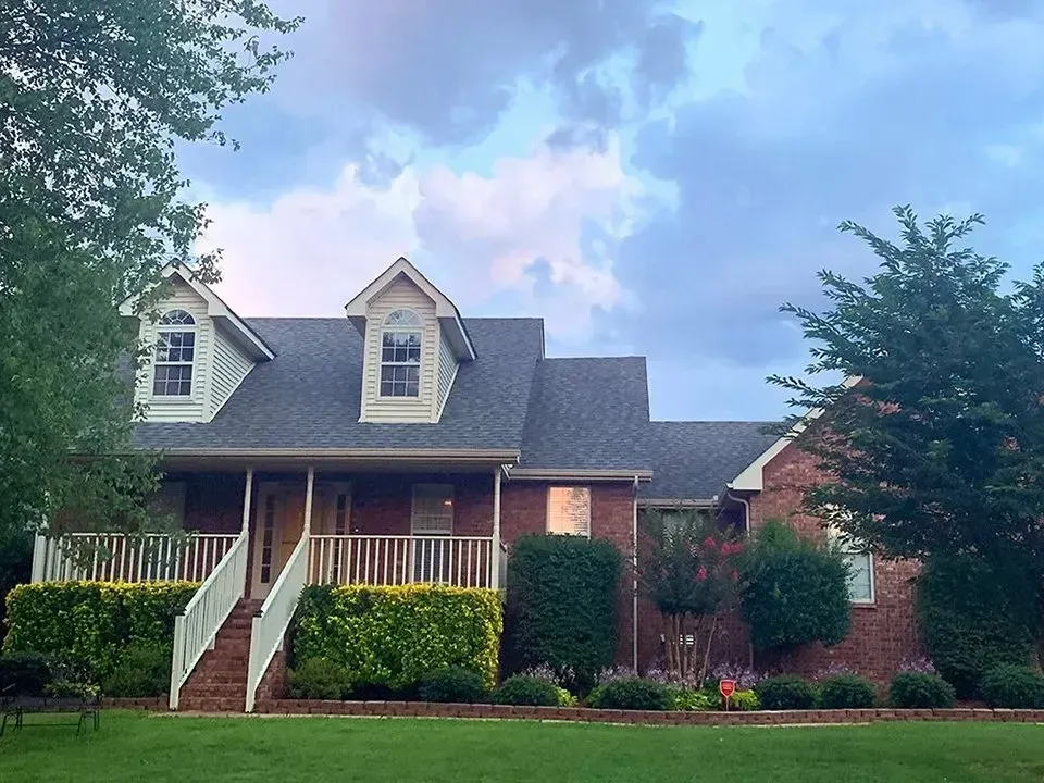 Brick house with a porch, dormers, and a lawn under a cloudy sky. Green shrubs and trees surround the house.