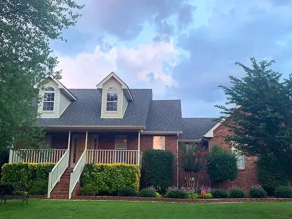 Brick house with porch, dormers, and a cloudy sky. Green lawn and bushes in front.