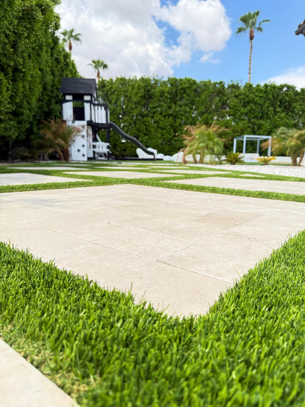 A backyard with concrete squares separated by grass, a playhouse, and a tall hedge.
