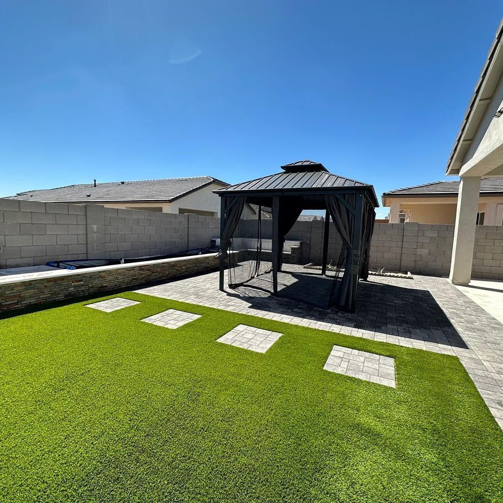 Gazebo in a backyard with artificial grass, stone path, and concrete block walls under a clear blue sky.
