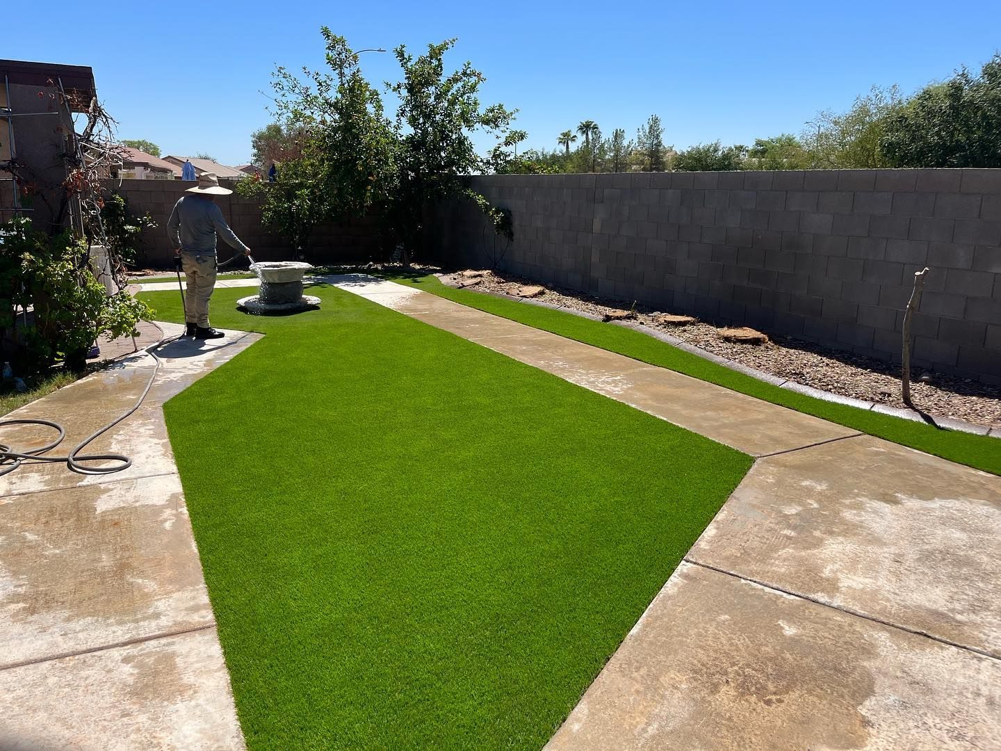 Person spraying a lush green artificial lawn in a backyard with a concrete path and walls.