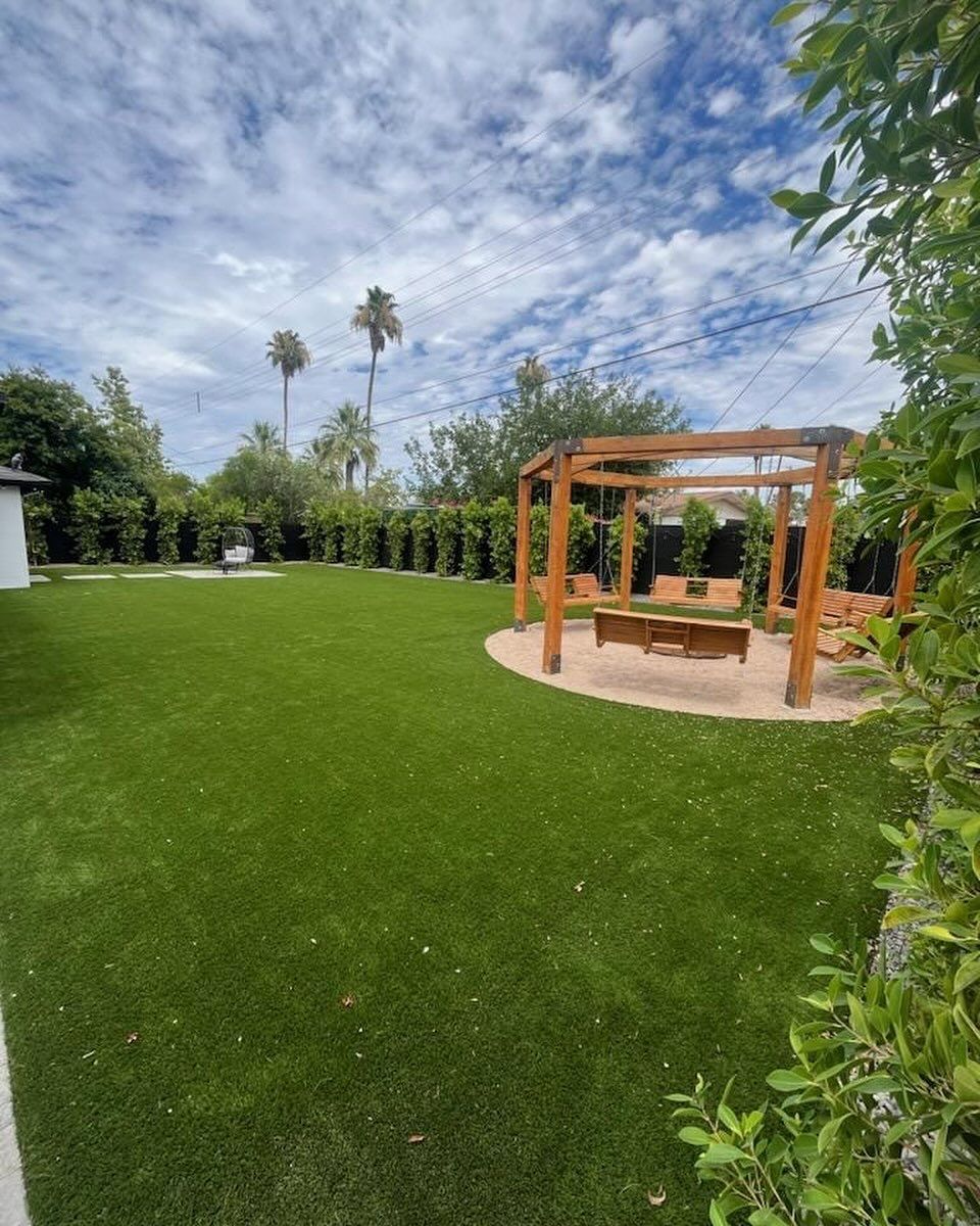 Backyard with green lawn, wooden swing, and trees under a cloudy sky.