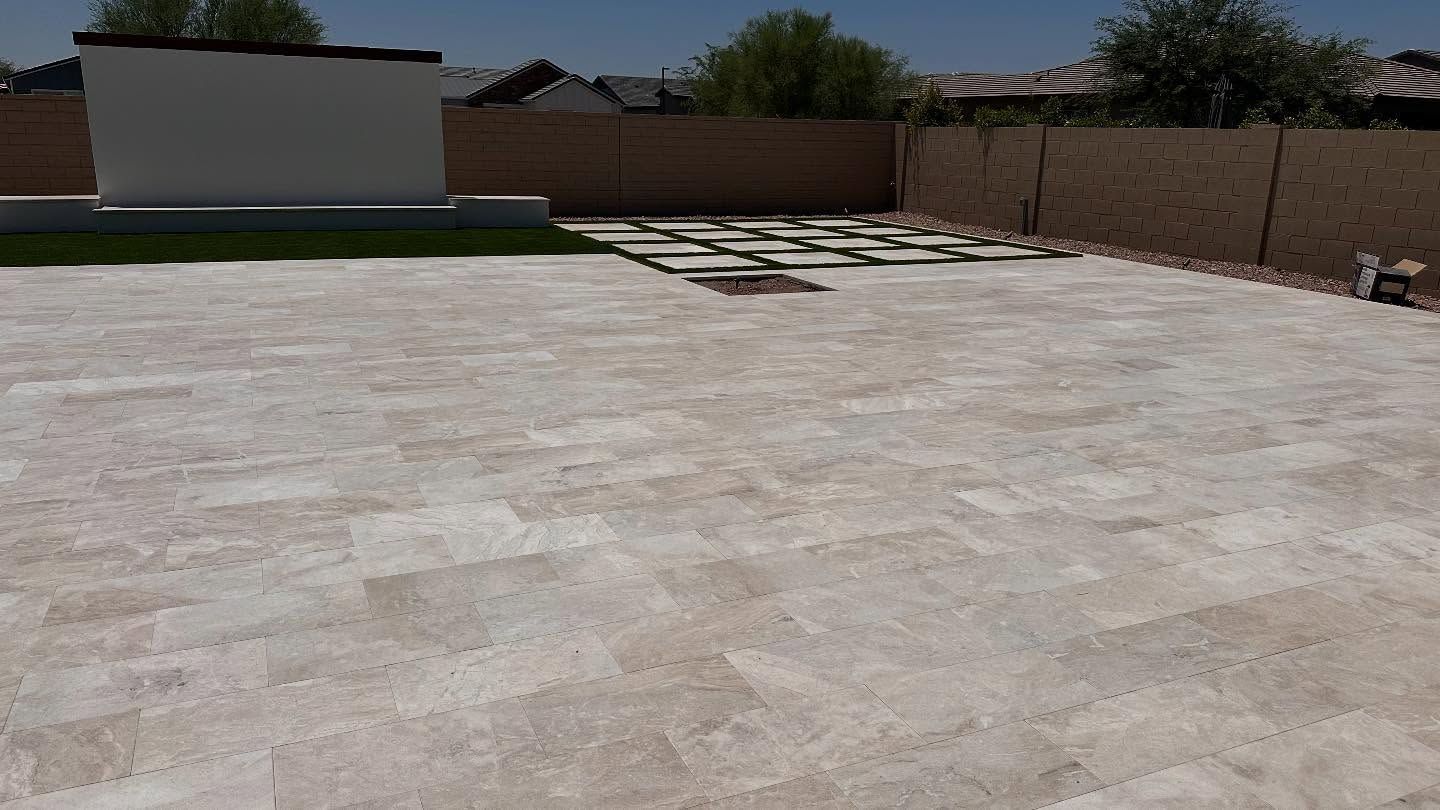 A large, light-colored stone patio in a backyard, with a green lawn section and a wall in the background.
