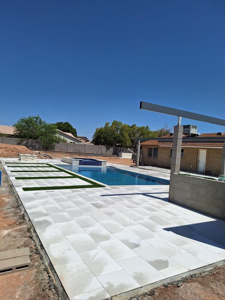 A rectangular pool with light gray tiles, blue water, and fake grass in a sunny outdoor setting.
