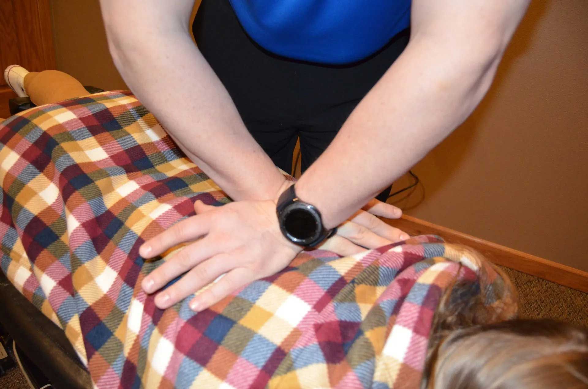 Hands on a person's back, likely performing chiropractic adjustment. Brown plaid blanket covers the person.