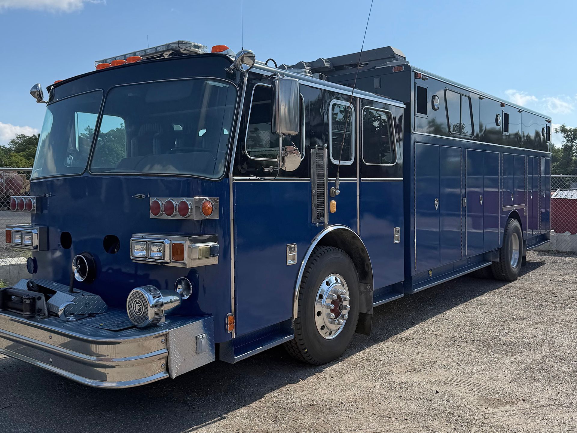 Blue fire truck parked on gravel, side view, with chrome details and roof-mounted equipment