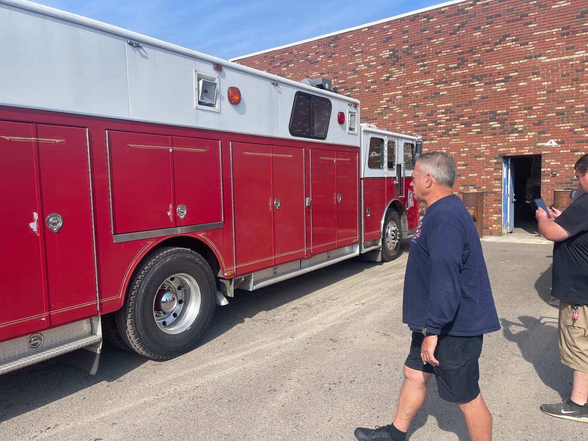Red fire truck parked beside a brick building, with two people walking nearby.