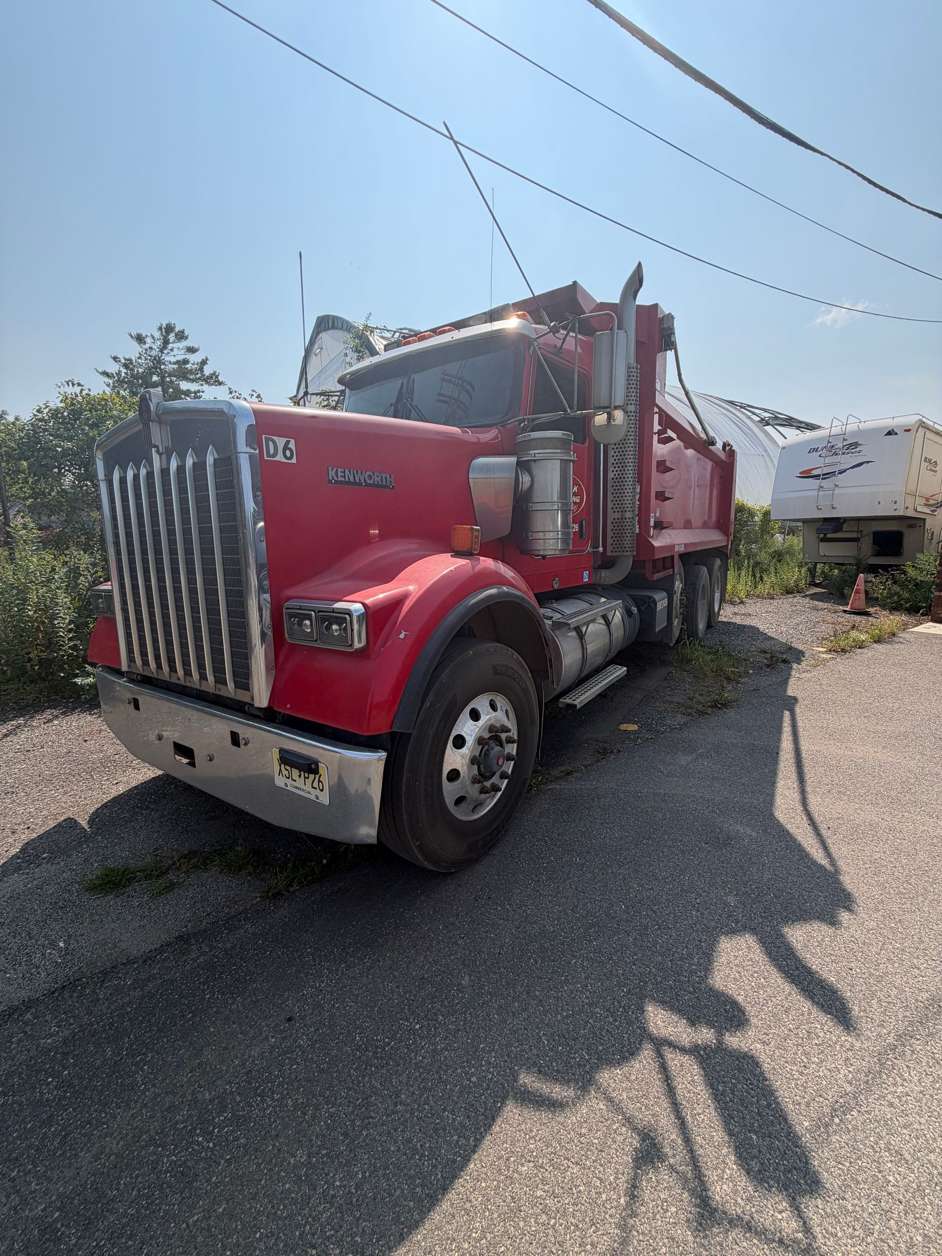 Red semi truck parked on gravel under a bright sky, with a white trailer in the background.