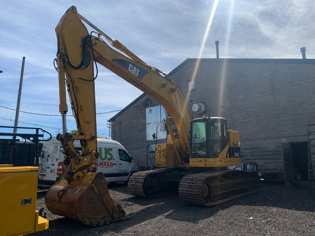 Yellow excavator on tracks beside a building in a gravel lot under bright sunlight.