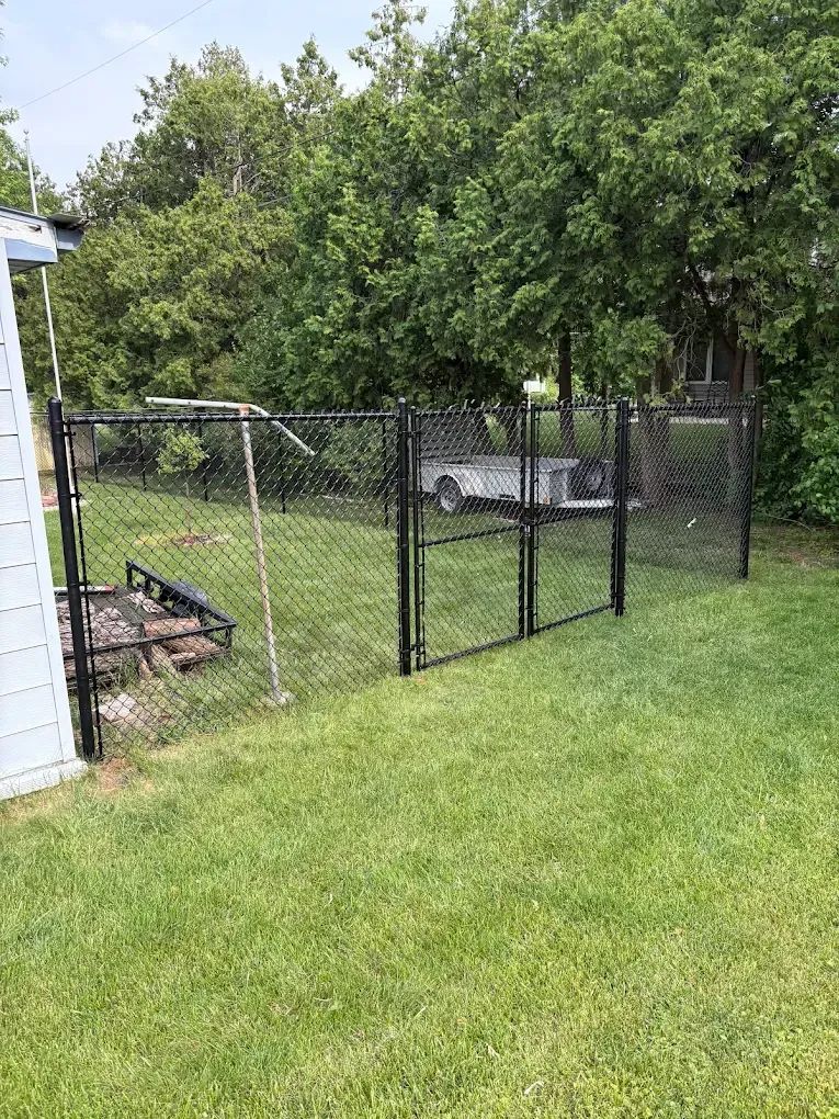 Black chain-link fence encloses a grassy yard; a gate is open, revealing a trailer and trees in the background.