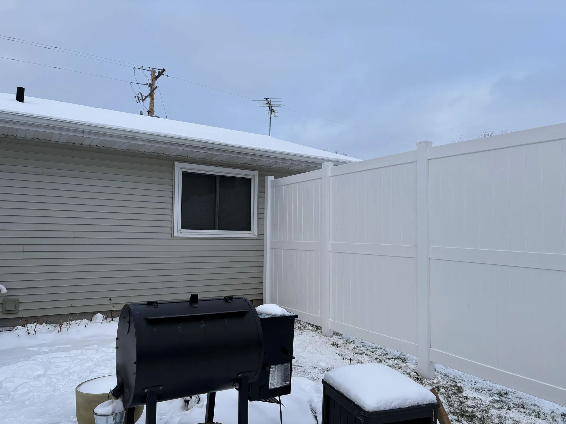 Snowy backyard scene with a black smoker grill, white fence, and a house under a cloudy sky.
