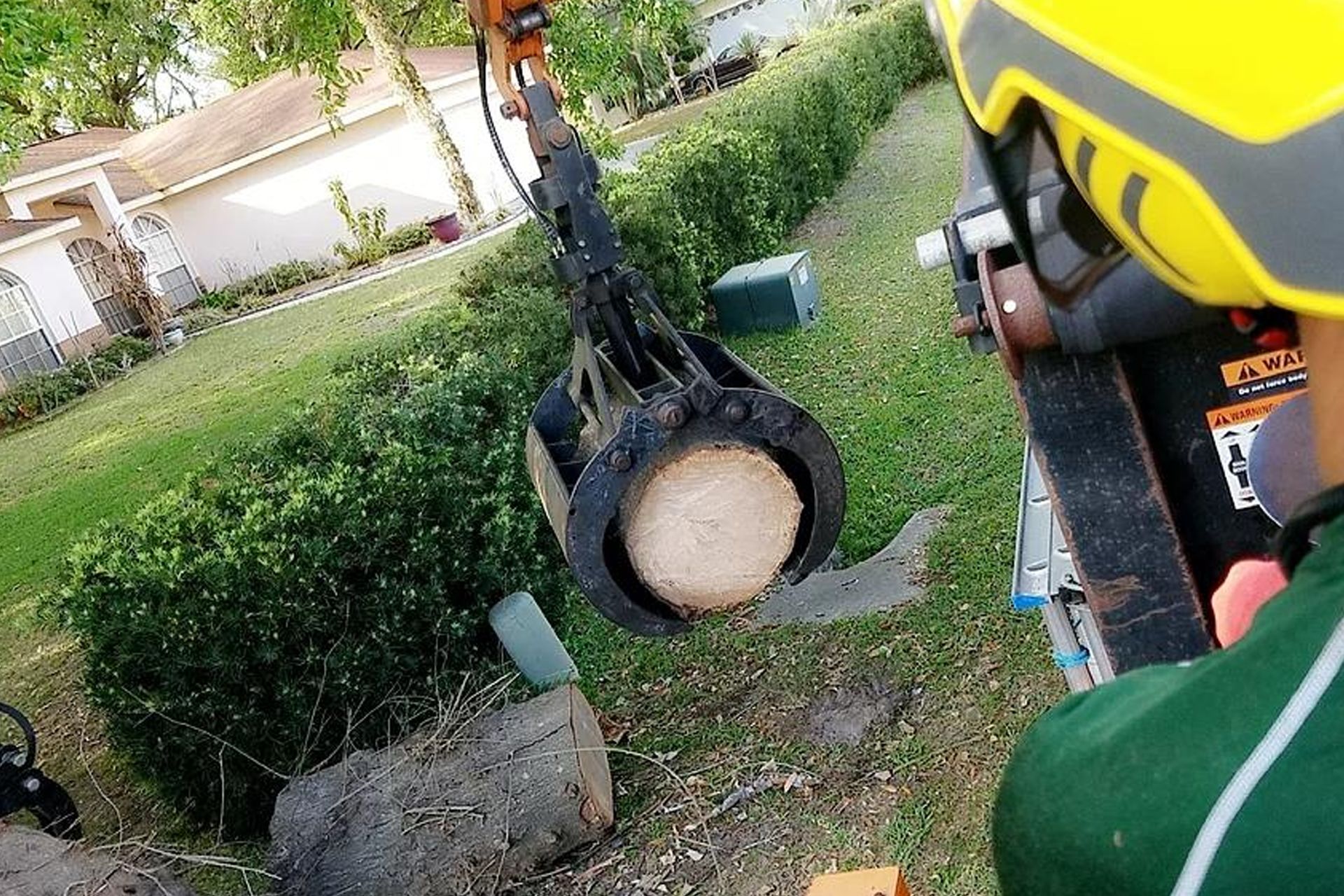 An arborist operating an excavator with a log in its claw, removing a tree trunk from a residential yard.