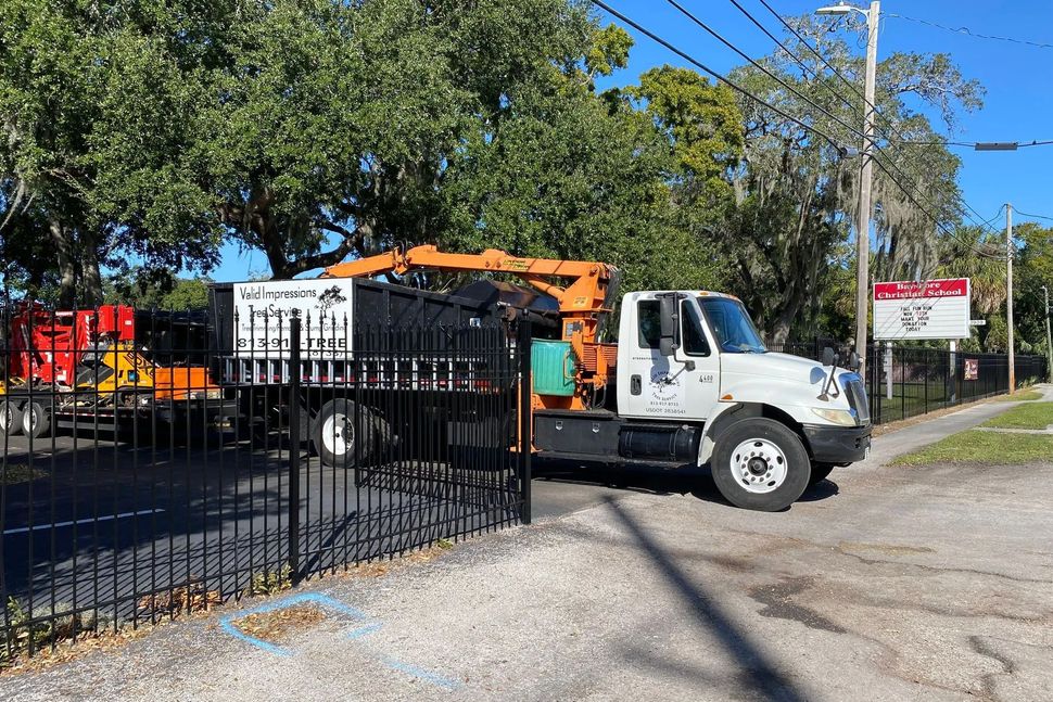 A white truck with a crane in front of a black metal fence; a tree, a trailer, and a sign are also visible.