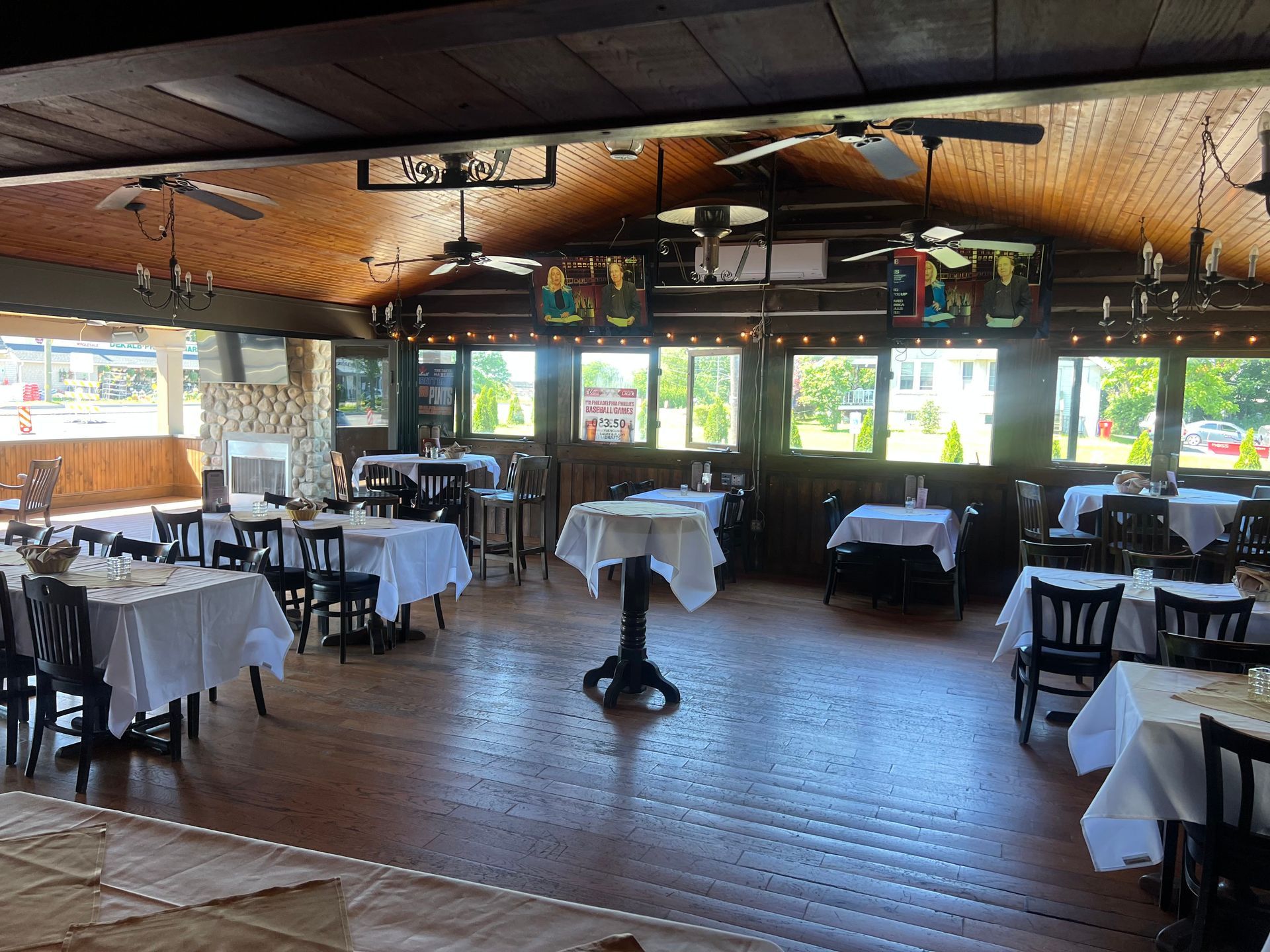 Empty dining room with white tablecloths, wooden floor, ceiling fans, and large windows overlooking greenery