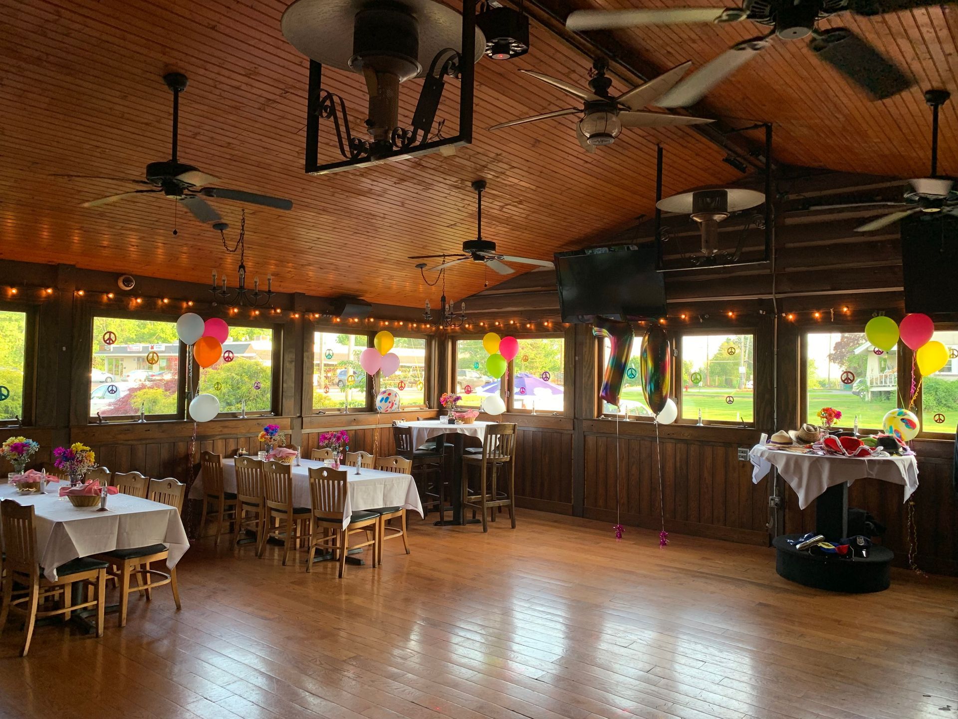 Decorated banquet hall with white tablecloths, balloons, wooden ceiling, and large windows overlooking greenery
