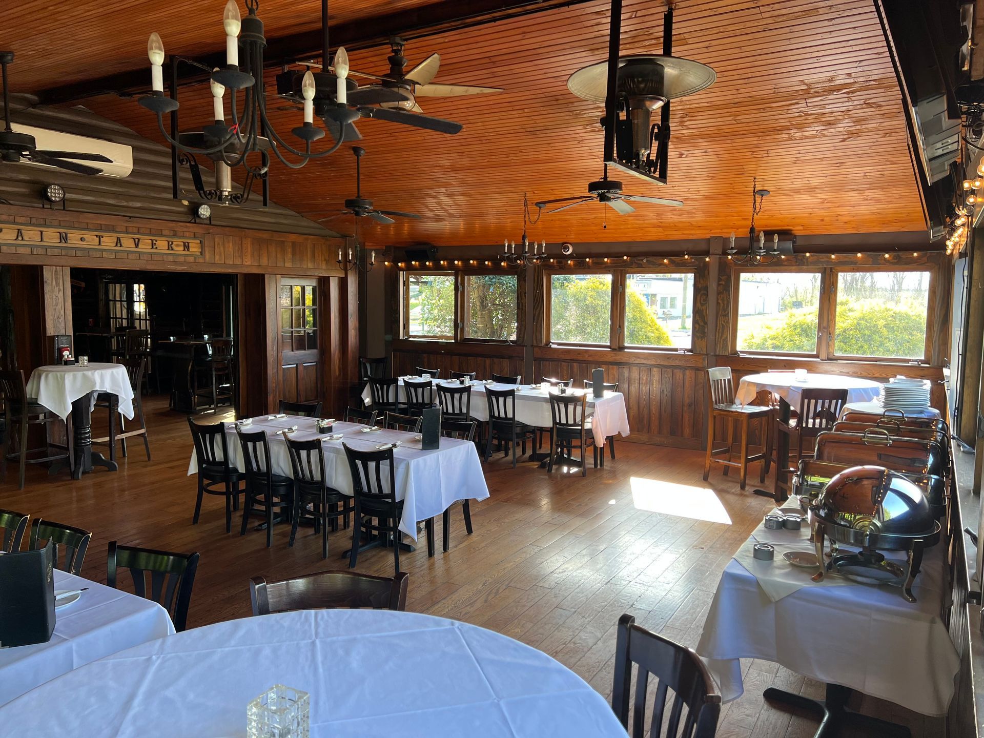 Empty rustic dining room with white tablecloths, wooden floor, and large windows overlooking greenery