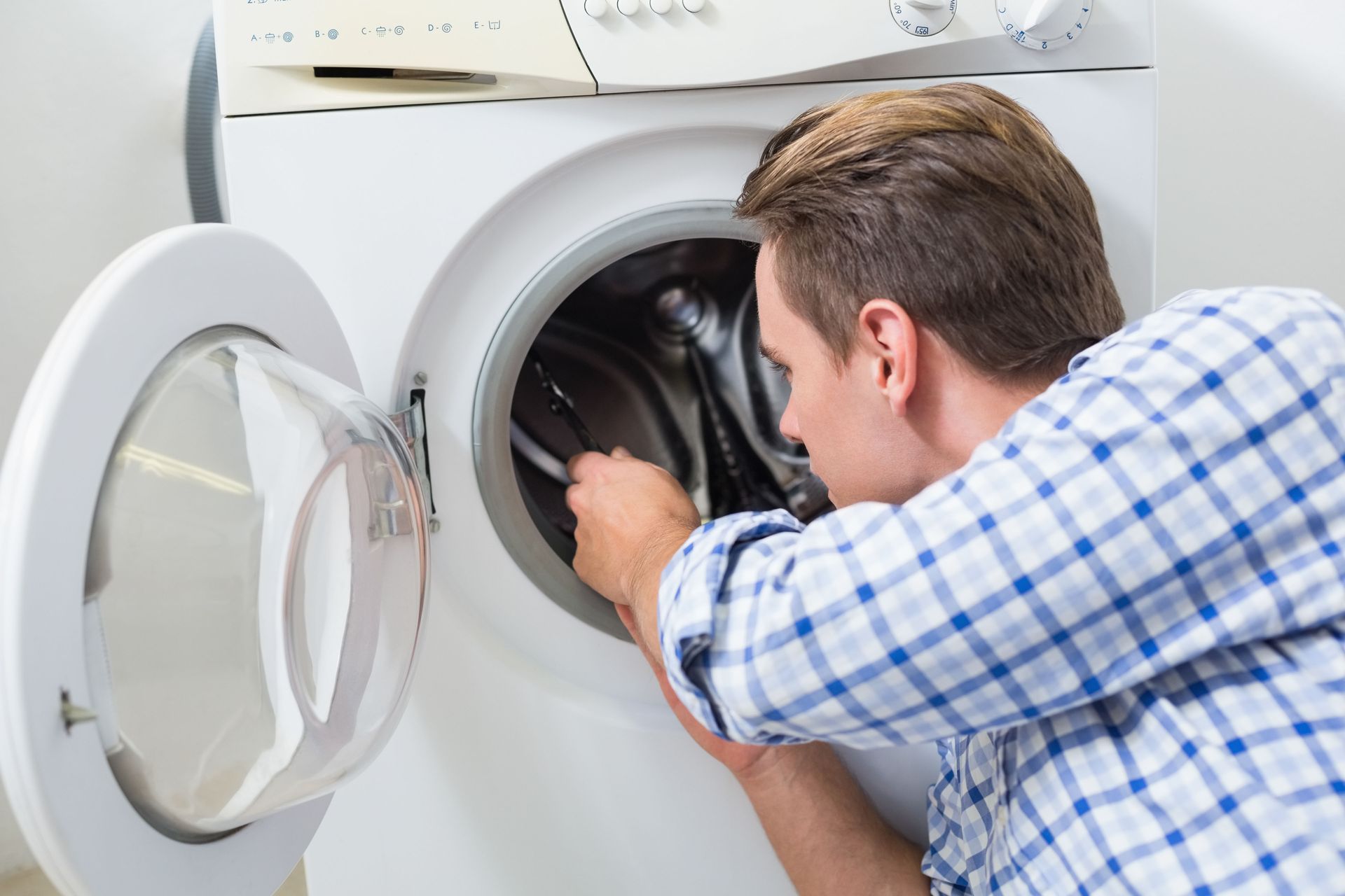 Man repairs washing machine. Open door, light interior, blue plaid shirt.