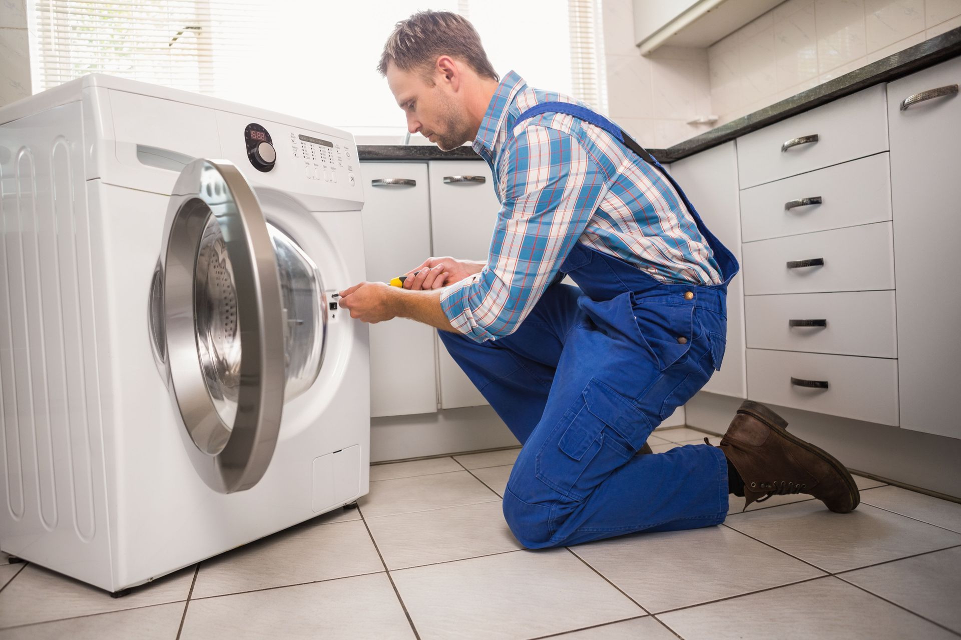 Man in blue overalls repairs a washing machine in a kitchen.