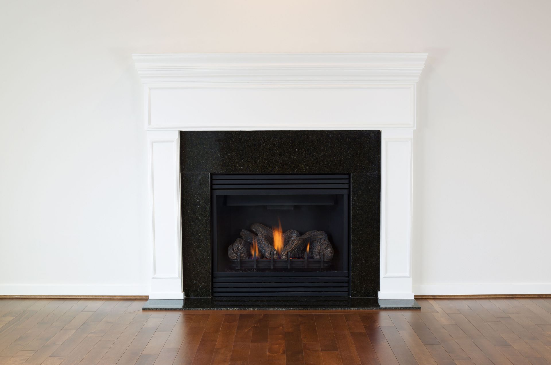 White mantel surrounding a black fireplace with flames, set against a white wall and hardwood floor.