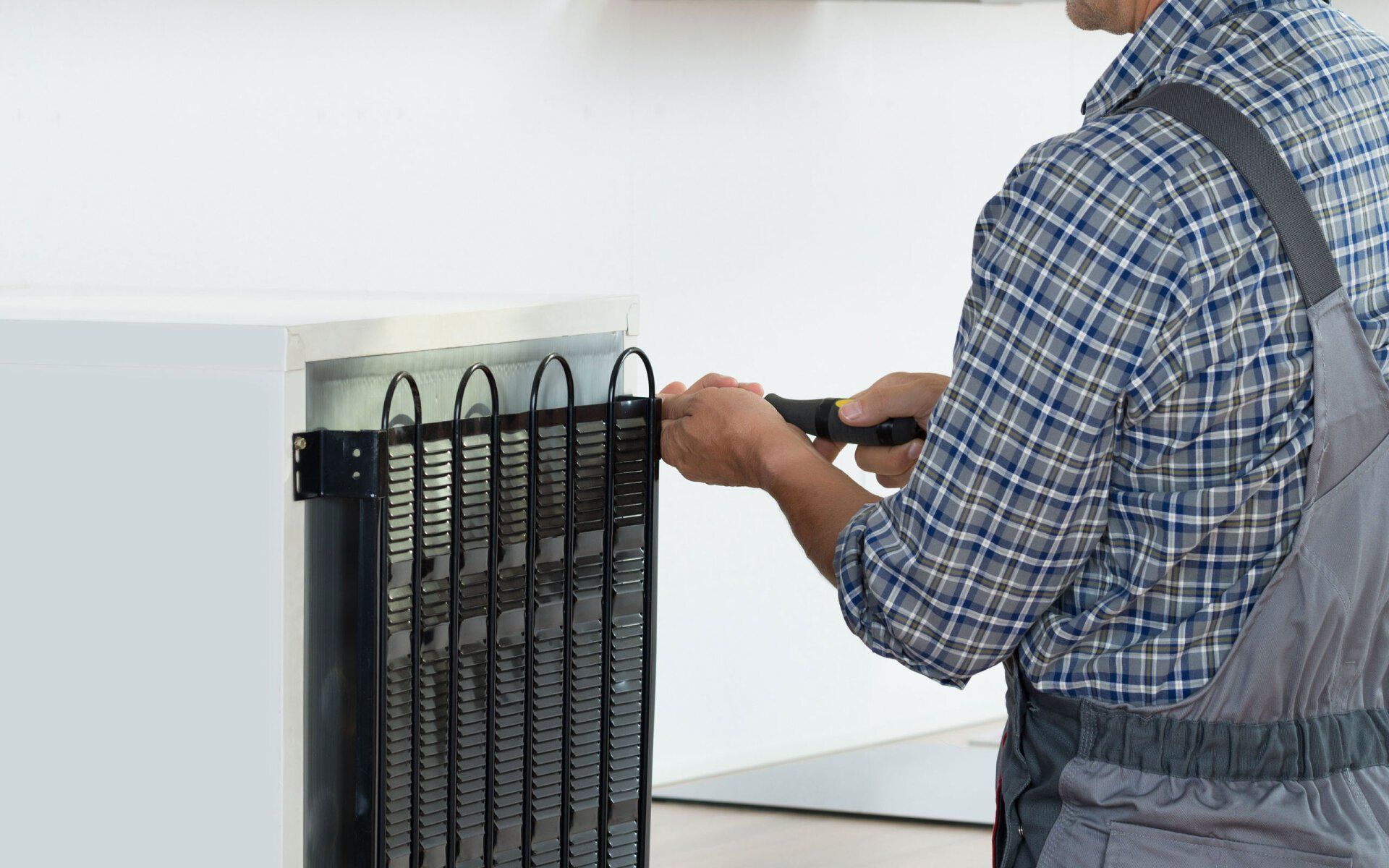 Person in overalls fixing the back of a refrigerator with a tool.