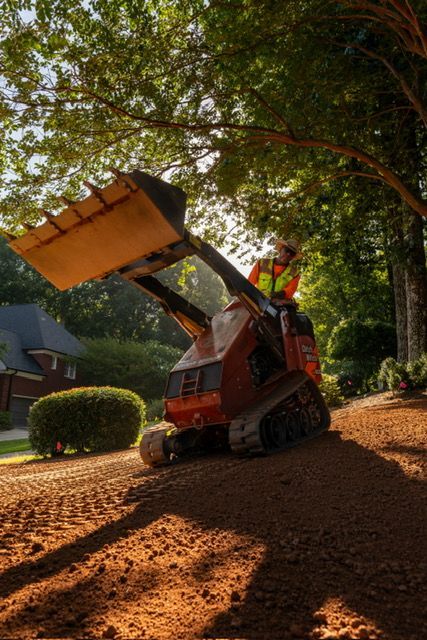 Orange skid-steer loader dumping mulch under trees in a landscaped yard