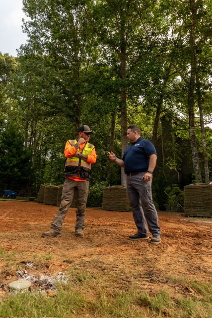 Two workers in safety gear talking on a muddy construction site near trees