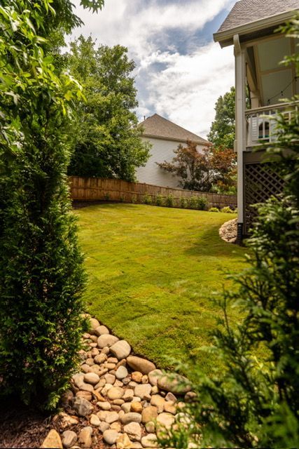 Backyard lawn framed by shrubs and rocks, with a fence and house under a cloudy sky