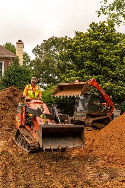 Orange skid steer moving dirt at a construction site with trees and a house in the background